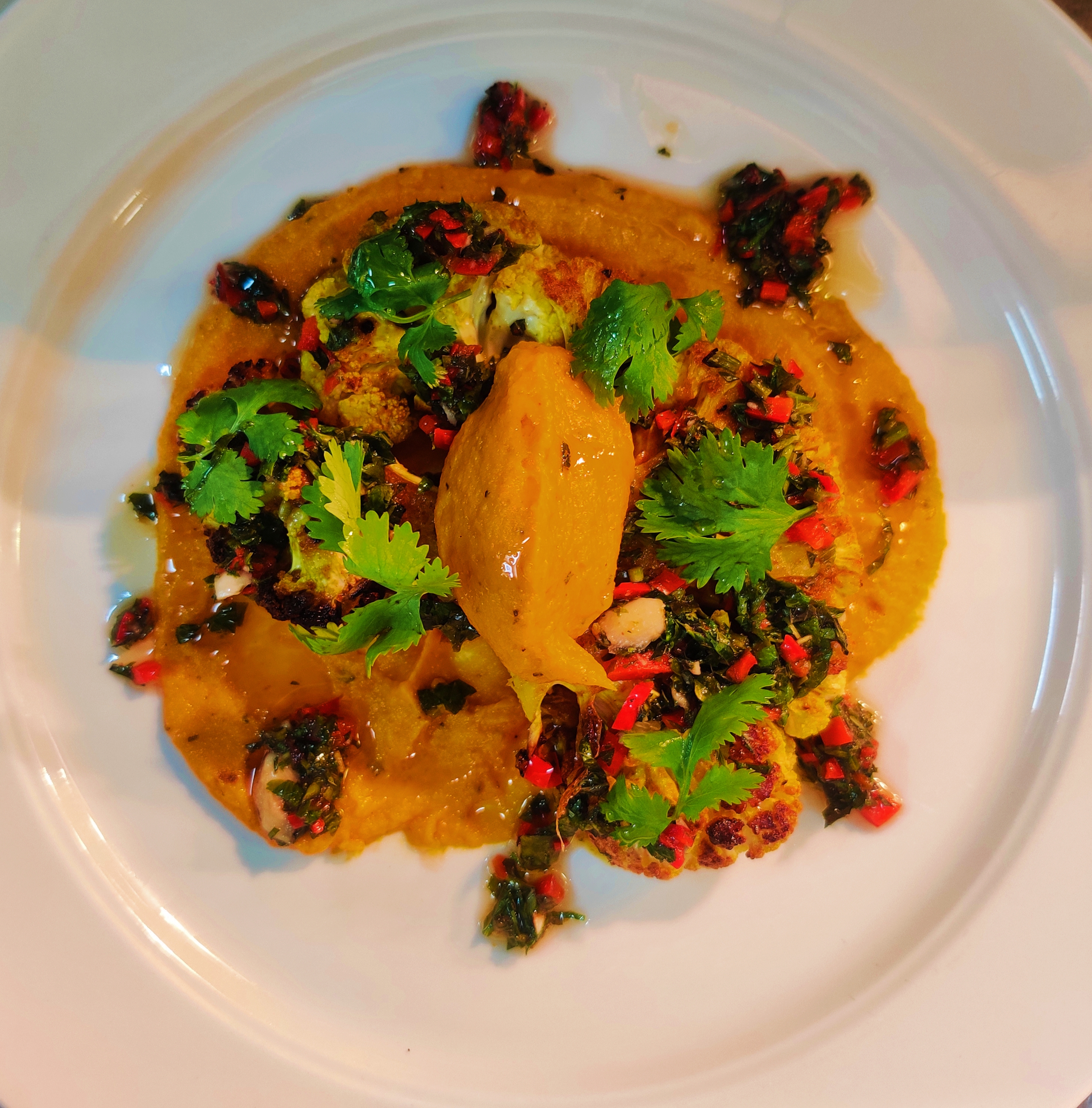 Charred cauliflower steak served with creamy carrot hummus and fresh chimichurri sauce on a rustic plate — a vibrant vegan dish from FeastyNet.net.