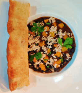 A bowl of Harissa Black Bean Ragout topped with roasted butternut squash, grated tofu, and fresh herbs, served with a golden focaccia slice.