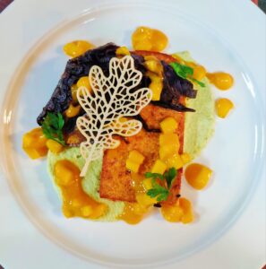 A beautifully plated vegan dish featuring marinated grilled tofu served on creamy edamame hummus, topped with roasted red cabbage, fresh mango salsa, and garnished with a crispy tuile and herbs on a white plate.