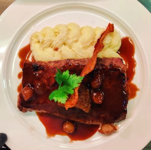 Grilled calf liver with burnt onion sauce, creamy mashed potatoes, crispy bacon, and parsley garnish on a white plate.