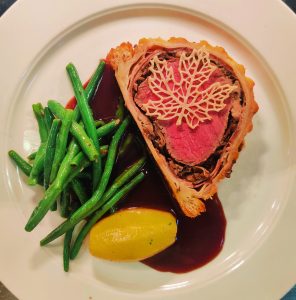 Slice of traditional beef Wellington with medium-rare beef, mushroom duxelles, potato fondant, sautéed garlic green beans, and red wine sauce on a white plate
